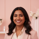 A smiling South Asian woman in her late 20s or early 30s, exuding genuine happiness and satisfaction, against a blush pink ba