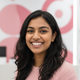 A smiling young adult South Asian woman, mid-shot, wearing a blush pink top, with a soft background in rose and white tones.